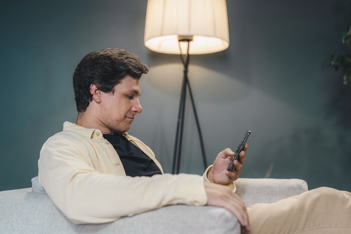Mężczyzna siedzący na kanapie w mieszkaniu wieczorem, patrzący na telefon