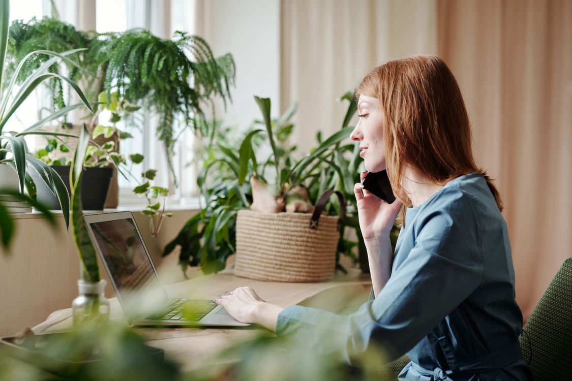 Kobieta rozmawia przez telefon przy laptopie w jasnym pokoju pełnym roślin